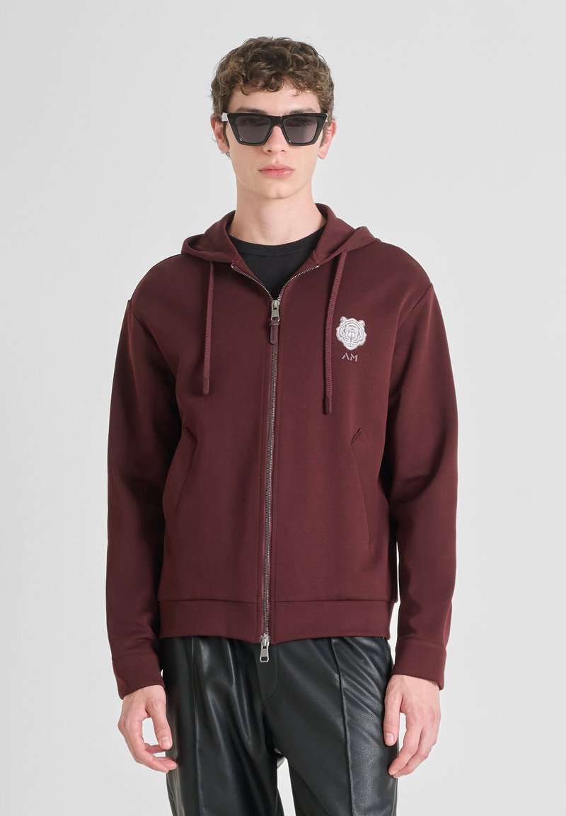 Felpa a zip color marsala con tasca frontale, coulisse e logo ricamato. Indossata sopra una maglietta nera e abbinata a pantaloni in pelle nera.