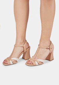 Sandales à talons hauts couleur nude et blanches avec un design à bride en T, des brides brillantes, un talon épais et un bout ouvert laissant apparaître les ongles des orteils.