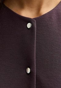 Ciemnofioletowa bluzka z okrągłym dekoltem, z dwoma widocznymi czarnymi guzikami z błyszczącymi srebrnymi akcentami na środku przodu.