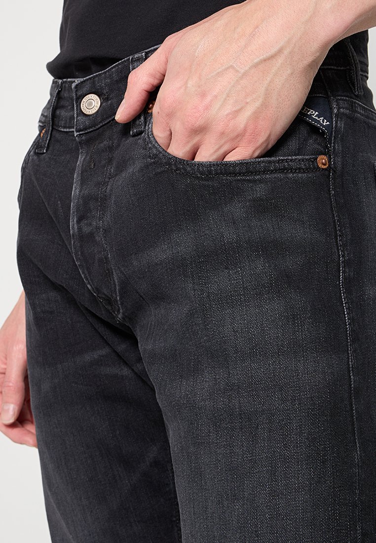 Close-up van een persoon in zwarte jeans met de rechterhand in de voorzak en een zichtbare riemlus.