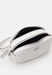 Bílá kožená taška s texturovaným povrchem, dvěma zipovými přihrádkami a plochým úchytem; na přední straně je malá kovová značka.