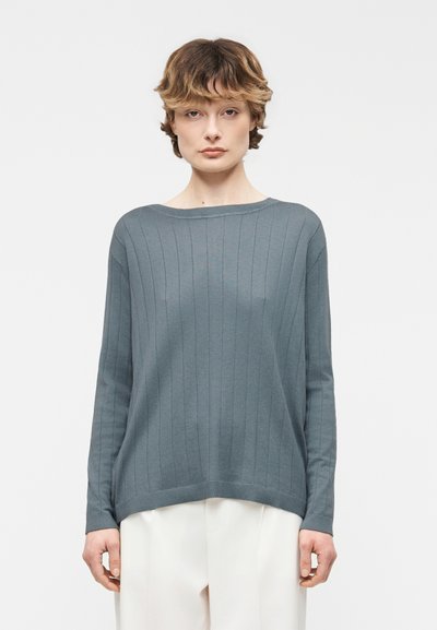 Jeune personne aux cheveux bruns courts portant un pull côtelé bleu-gris à manches longues et un pantalon blanc, debout devant un fond uni.