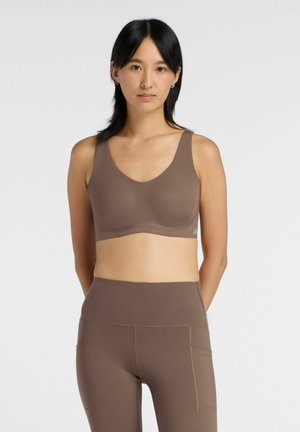 Vrouw met zwart haar die een bruine sportbeha en bijpassende high-waist legging draagt, staand tegen een effen witte achtergrond.