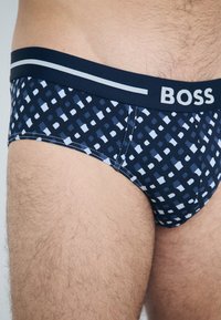 Tamsiai mėlynos bokseriai su šviesiai mėlynu geometriniu raštu. Platus juosmuo su užrašu "BOSS" kontrastine balta spalva. Lygus paviršius.