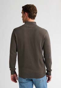 Maglione lavorato a maglia di un verde scuro con colletto alto, motivo testurizzato e polsini a coste, indossato sopra jeans azzurri chiari, che mostrano la vista posteriore.