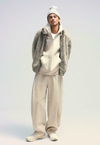 Jachetă fleece crem cu fermoar, cu o textură moale, asociată cu un hanorac asortat și pantaloni de trening cu picioare largi. Căciulă gri deschis. Tenisi albi.