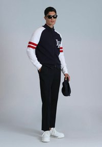 Marineblaue und weiße Jacke mit Reißverschluss und roten Streifen, schwarze taillierte Hose und weiße Sneaker. Das Model trägt Sonnenbrillen und hält eine marineblaue Mütze.