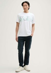 Hvid bomulds t-shirt med grønt og gråt grafikdesign henover brystet, parret med mørkeblå denimjeans og hvide sneakers.