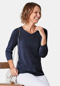 Dunkelblauer Strickpullover mit V-Ausschnitt, dreiviertellangen Ärmeln und Spitzenakzenten am Ausschnitt, kombiniert mit weißen Hosen und einer hellen Handtasche.
