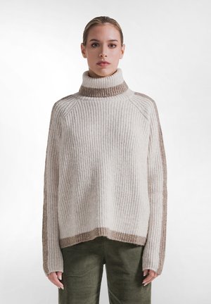Kvinde med lys hud og sammenbundet hår iført en beige og brun ribstrikket rullekravesweater og olivengrønne kordfløjlsbukser.
