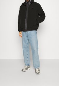 Black zip-up jacket with high collar, blue jeans, and gray sneakers. Features include a subtle logo and a mix of smooth and textured fabrics.