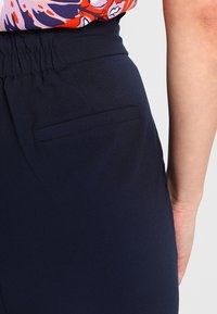 Námořnické modré kalhoty s hladkou texturou a detailem zadní kapsy, vyrobené z lehkého, splývavého materiálu. Viditelný elastický pas.