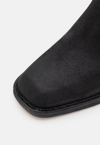 Gros plan sur l'avant d'une botte en daim noir avec une forme carrée et des coutures visibles le long du bord de la semelle sur un fond blanc.