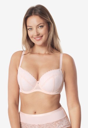 Kobieta z długimi blond włosami uśmiechająca się, ubrana w jasnoróżowy koronkowy biustonosz i pasujące koronkowe majtki z wysokim stanem na białym tle.