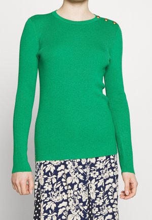 Femme portant un pull côtelé vert à manches longues avec trois boutons dorés à l'épaule et une jupe bleue à motifs floraux blancs.