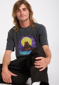 Jeune homme aux cheveux longs assis en tailleur, portant un T-shirt noir avec un motif coloré de volcan et un pantalon noir sur un fond uni.