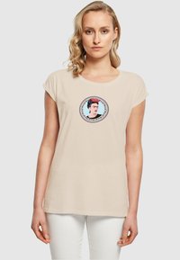 Beige bomulds T-shirt med korte ærmer, der har et farverigt grafik af en kvinde i en cirkulær ramme, med indviklede mønstre omkring kanten.