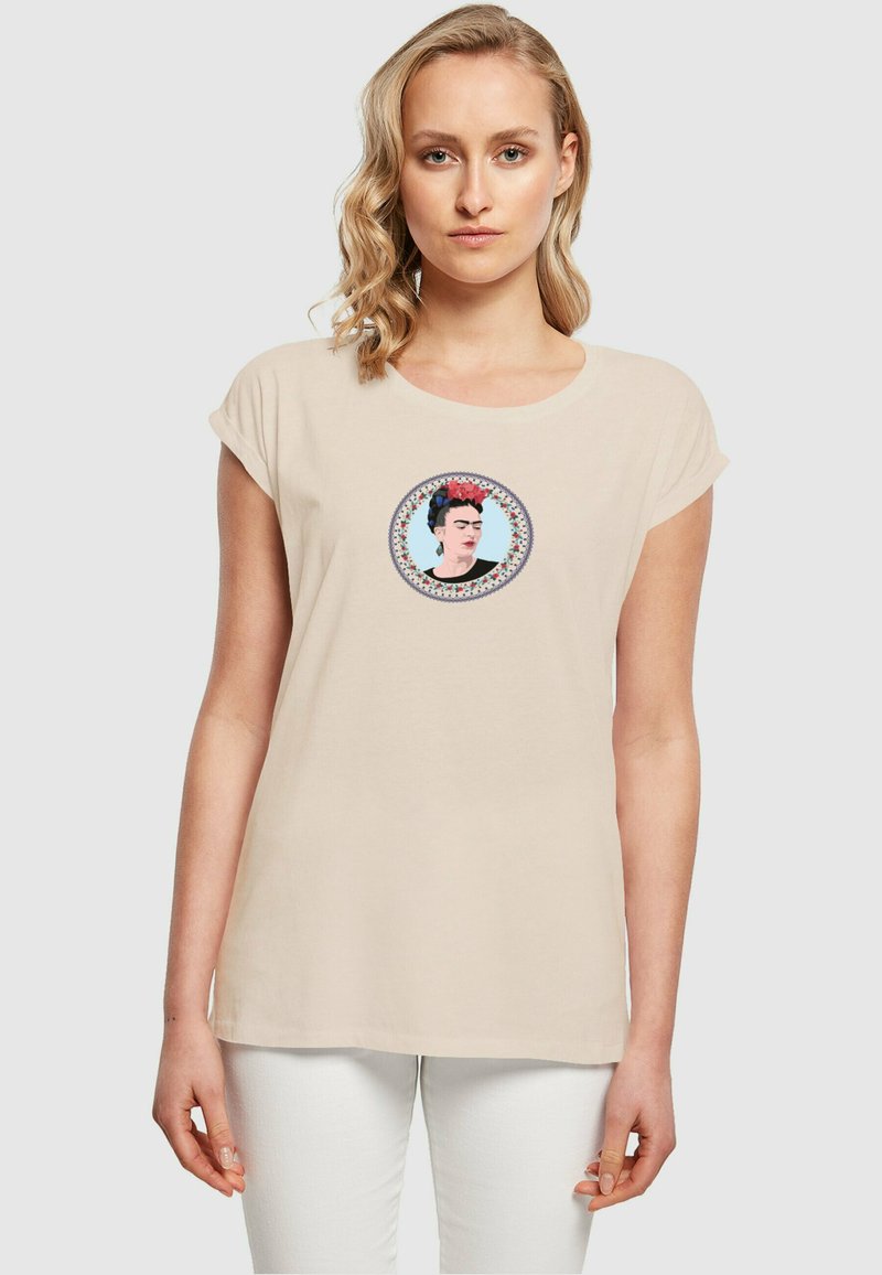 Beige bomulds T-shirt med korte ærmer, der har et farverigt grafik af en kvinde i en cirkulær ramme, med indviklede mønstre omkring kanten.