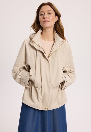 Kobieta w beżowej lekkiej kurtce z zamkiem z przodu i rękami w kieszeniach, w połączeniu z niebieską spódnicą na jednolitym tle.