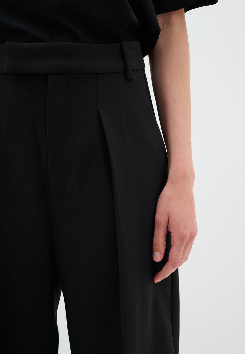 Schwarze, maßgeschneiderte Hose mit Falten, einer glatten Textur und einer breiten Gürtelschlaufe an der Taille. Die Hand zeigt eine entspannte Positionierung auf dem Stoff.