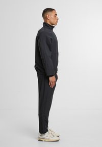 Černý sportovní teplákový komplet s vysokým límcem a odpovídajícími kalhotami, vyrobený z lehkého materiálu s hladkou texturou. Na nohou bílé tenisky.