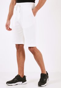 Hvide sweatshorts med ribbet tekstur, elastisk talje og snøre, med sidelommer og knælængde. Båret med sorte sneakers.