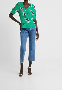 Blusa corta de manga corta en verde con flores, con escote en V y detalles de pliegues en el frente, combinada con jeans cortos de tono azul claro y tacones de tiras beige.
