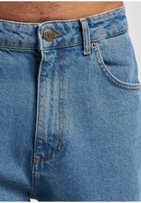Vaqueros de mezclilla de color azul claro con cierre de botón, diseño de cinco bolsillos, costuras de color cobre y ligeras marcas de desgaste en la tela.