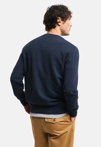 Sudadera azul marino de manga larga, cuello redondo y puños acanalados, combinada con pantalones beige. Textura suave, diseño minimalista.