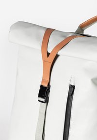 Weißer Rolltop-Rucksack aus strapazierfähigem Material. Verfügt über einen hellbraunen Lederriemen mit einem schwarzen Clip und eine seitliche Reißverschlusstasche.
