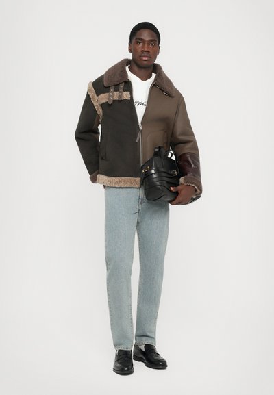 Mann trägt eine zweifarbige Lammfelljacke, hellblaue Jeans, schwarze Slipper und hält eine schwarze Lederhandtasche vor einem weißen Hintergrund.