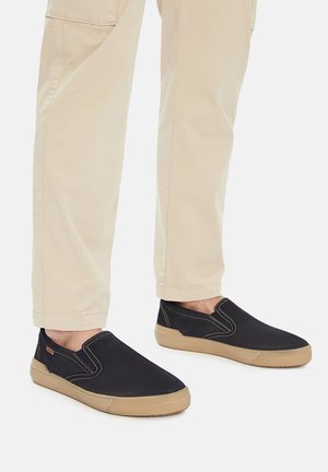 Zapatos negros sin cordones con suelas de goma beige, con costuras en contraste y un diseño minimalista, usados con pantalones beige claro.