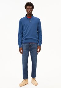Pullover con zip blu, strato interno rosso, abbinato a jeans in denim di lavaggio medio e scarpe morbide beige, caratterizzato da una vestibilità rilassata e una superficie liscia.
