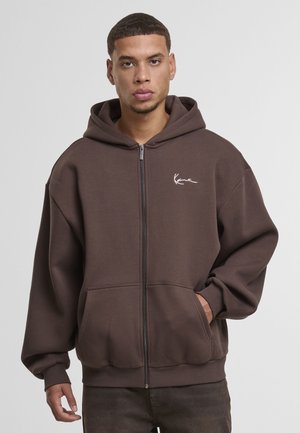 Brauner Kapuzenpullover mit Reißverschluss aus dickem Stoff, ausgestattet mit einem Kordelzug an der Kapuze, zwei Vordertaschen und einem kleinen weißen Logo auf der linken Brust.