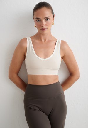 Femme aux cheveux tirés en arrière, portant des boucles d'oreilles créoles dorées, un crop top blanc sans manches et un legging taille haute foncé, devant un mur blanc.