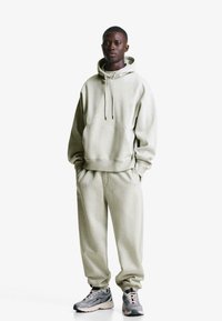 Világosszürke kapucnis pulóver elülső zsebbel, megegyező színű melegítőnadrággal párosítva. Az anyag puha tapintású; laza fazonú, bordás mandzsettákkal rendelkezik.