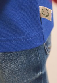 Blue shirt hem with a small fabric tag featuring a green smiling face icon above blue denim jeans.