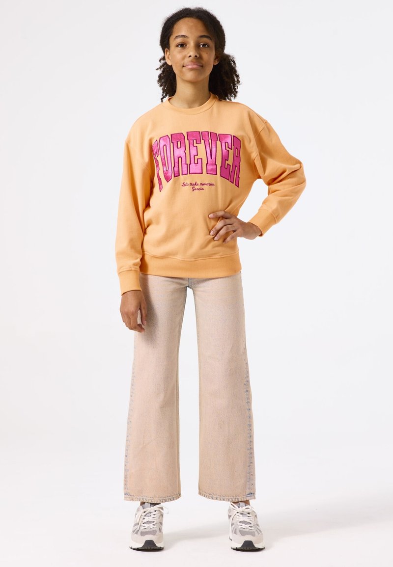 Sudadera naranja con "FOREVER" en rosa, combinada con pantalones vaqueros de pierna ancha de color claro y zapatillas blancas con acentos grises.