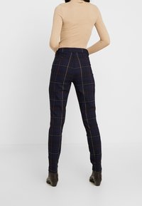 Pantalons à carreaux navy avec des motifs à checks jaunes et rouges, style ajusté, texture lisse et taille mi-haute. Des bottines marron sont portées.