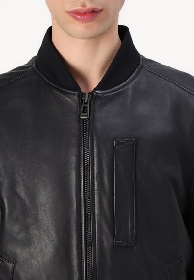Veste bombardier en cuir noir avec un col côtelé, une fermeture éclair à l'avant et un détail de patch rectangulaire sur le côté gauche. Texture lisse.