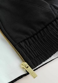 Prenda negra con cremallera y tirador dorado, fabricada con tela elástica negra, superpuesta sobre un textil blanco.