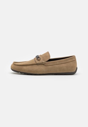 Mocassins en daim beige avec un bout arrondi, des détails cousus et un petit ornement métallique décoratif à l'avant. Semelle en caoutchouc noir flexible.