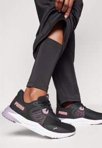 Zwarte Puma-sneakers met roze accenten, bovenwerk van mesh, witte zool en een patroonachtige rubberen loopzool, gedragen met zwarte sportbroek.
