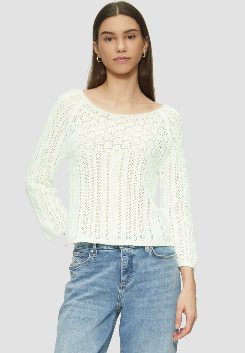 Weißer gestrickter Pullover mit einem runden Halsauschnitt, langen Ärmeln und einem Lochmuster. Mit hellblauen Denim-Jeans kombiniert.