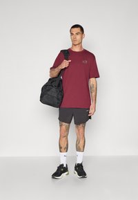 Rødbrun kortærmet T-shirt med et lille logo, parret med grå shorts. Sort træningspose, sorte sneakers med grønne detaljer og hvide sokker.