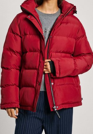 Personne portant une doudoune rouge vif par-dessus une chemise grise et un pantalon bleu marine rayé, en train de fermer partiellement la fermeture éclair de la doudoune.