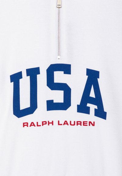 Bela majica s polovičnim zadrgo in ovratnikom, ki ima velik modri napis "USA" ter "RALPH LAUREN" v rdeči barvi pod njim. Mehak tekstura tkanine.