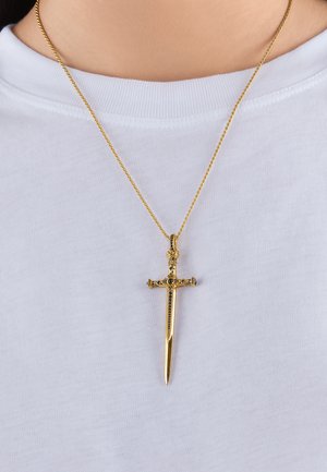 Goldener schwertförmiger Anhänger mit schwarzen Edelsteindetails hängt an einer feinen Kette vor einem weißen Hemd.