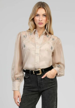 Femme blonde portant un chemisier beige transparent brodé de fleurs avec des manches bouffantes, une ceinture noire avec boucle dorée, et un jean noir taille haute.