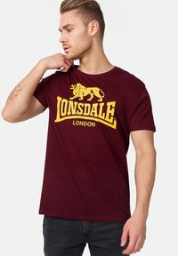 Burgundi színű póló sárga oroszlán logóval és "LONSDALE LONDON" felirattal. Rövid ujjú és kerek nyakú. Pamut anyag, laza fazon.